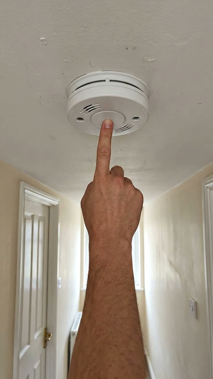 A finger pressing the test button on a smoke alarm on a hallway ceiling